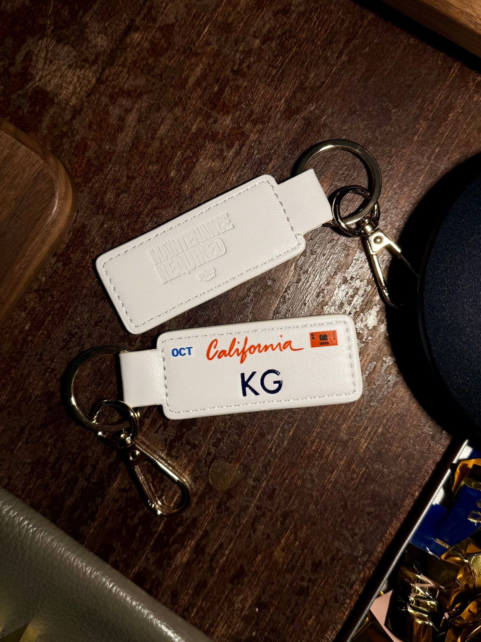 White_License_Plate_Keychains_KU_Blue_Foil_Maintenance_Required_Prime_Video Two white vegan leather license plate keychains from Prime Video’s Maintenance Required premiere, one embossed with KU in dark blue foil and the other showing the debossed “Maintenance Required” and “Prime Video” logos.